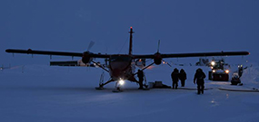 Photo of a small aircraft on the snowy ice cap in the evening, with people and a car to the right of the plane.